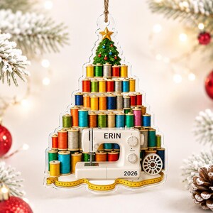 May include: A clear, tree-shaped Christmas ornament featuring a stack of colorful sewing thread spools, a white sewing machine labeled ERIN with the year 2026, a small green tree topper with a gold star, and a yellow measuring tape at the base.