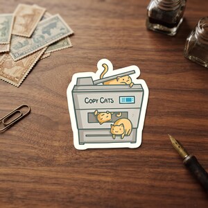 May include: A die-cut sticker featuring a gray office photocopier with three orange cats interacting with it. One cat lies on the scanner bed, while two others are positioned within the paper tray area. The text Copy Cats is printed in black.