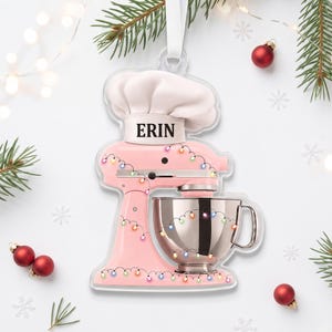 May include: A clear acrylic Christmas ornament shaped like a pink stand mixer with a white chef hat on top. The hat features the name ERIN in black text. The mixer is decorated with colorful string lights and includes a metallic silver mixing bowl.