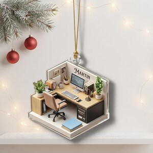 May include: A flat, acrylic hanging ornament featuring a miniature office scene with a desk, computer, chair, and potted plants. The wall of the office space displays the name MARTIN in black lettering. The ornament hangs from a gold-colored cord.