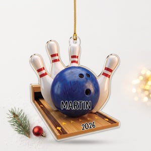 May include: A hanging ornament featuring a blue bowling ball with the name MARTIN printed on it, positioned on a wooden bowling lane with white pins and red stripes. The lane displays the year 2026 in black text, suspended by a gold cord.