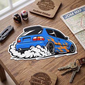 May include: A die-cut vinyl sticker featuring a blue hatchback car with orange flame graphics, black wheels, and a rear spoiler, depicted drifting with white tire smoke. The sticker is displayed on a wooden surface alongside two miniature helmets.
