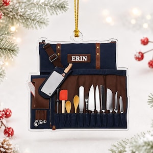 May include: A flat, clear acrylic ornament shaped like a chef's knife roll bag. The design features a navy blue and brown fabric texture with various kitchen tools, including knives, spoons, and a spatula. The name ERIN is printed in white.