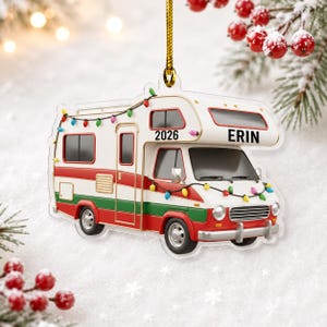 May include: A flat, acrylic Christmas tree ornament shaped like a vintage camper van. The vehicle is white with red and green stripes, decorated with colorful string lights. It features the text 2026 and the name ERIN, hanging by a gold cord.