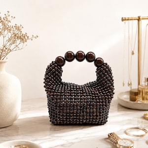 Op de afbeelding: Een kleine, donkerbruine handtas met kralen en een afgeronde handgreep van grote, bijpassende kralen. De tas staat op een marmeren oppervlak, met sieraden en een horloge in de buurt. Een vaas met gedroogde bloemen staat op de achtergrond.