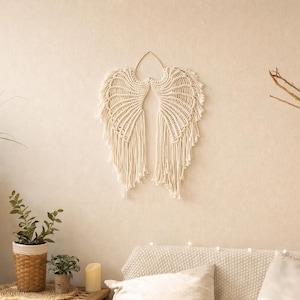 Op de afbeelding: Crèmekleurige macramé engelenvleugels wanddecoratie. De vleugels zijn ingewikkeld geknoopt en gefranjerd, waardoor een textuur met gevederde uitstraling ontstaat. Het kunstwerk hangt aan een muur met neutrale tinten en voegt een vleugje bohemien stijl toe aan de ruimte.