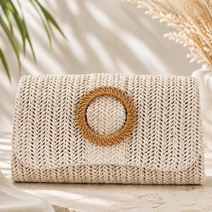 Könnte beinhalten: Beige gewebte Clutch mit einem kreisförmigen, braunen Griff. Die Clutch hat ein strukturiertes Fischgrätmuster und einen Klappenverschluss. Der Griff besteht aus gewebtem Material. Die Tasche ist rechteckig und scheint aus Naturfasern zu bestehen.