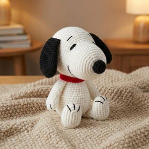 Op de afbeelding: Een handgemaakt Snoopy-pluche speelgoed, gehaakt. Het speelgoed is voornamelijk wit met zwarte oren, een zwarte neus en een rode kraag. Het pluche zit op een beige deken.