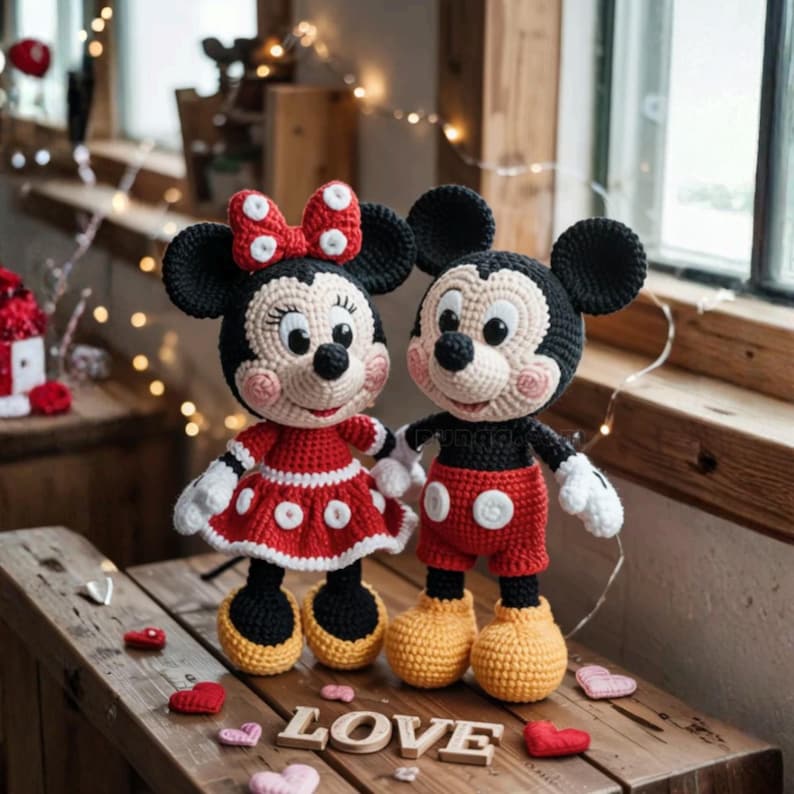 May include: Crocheted Mickey and Minnie Mouse dolls. Minnie wears a red dress with white polka dots and a matching bow. Mickey wears red shorts with white buttons. Both dolls have black ears and are standing on a wooden surface with the word "LOVE".