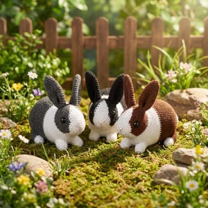 Może przedstawiać: Trzy szydełkowe króliki w ogrodzie. Jeden jest szaro-biały, drugi czarno-biały, a trzeci brązowo-biały. Siedzą na zielonym mchu, z drewnianym płotem w tle. Jasna i kolorowa scena.