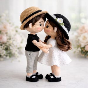 Puede incluir: Muñecos de ganchillo de una pareja abrazándose. El muñeco masculino viste una camisa negra, pantalones grises y un sombrero color canela con una banda negra. La muñeca femenina lleva un vestido blanco y un sombrero negro con una flor blanca. Ambos muñecos tienen zapatos negros.