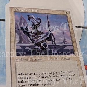 May include: A Magic: The Gathering card titled "Esper Sentinel." The card features an illustration of a humanoid figure in armor with a spear, set against a futuristic cityscape. Text includes card type, rules, and artist credit.