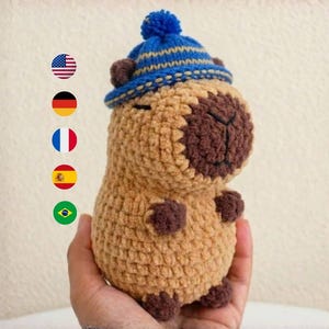 Peut inclure: Une peluche capybara crochetée, dans les tons de beige et de marron, portant un bonnet tricoté bleu et beige avec un pompon. L'animal en peluche fait main a de petits pieds et oreilles marron. La peluche est tenue dans une main.