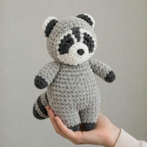 May include: A small, handmade crochet raccoon plush toy featuring a light gray body, a white face, and dark gray patches around the eyes. The raccoon has dark gray ears, paws, and a striped tail, all crafted with a soft, textured yarn finish.