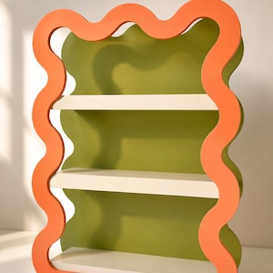 May include: A modern, three-shelf bookcase with a wavy orange frame and a green interior. The shelves are a neutral off-white color. The bookcase is approximately 30 inches tall and 20 inches wide.