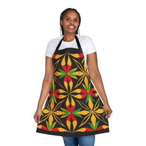 Op de afbeelding: Zwarte schort met een kleurrijk bloemmotief in rood, geel en groen. De schort heeft een zwarte nekband en strikken. De schort wordt gedragen door een persoon in een wit t-shirt en een blauwe spijkerbroek.