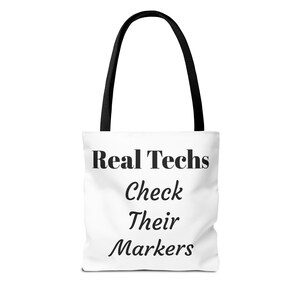 May include: White tote bag with black handles featuring the text "Real Techs Check Their Markers" in black. The bag is designed for carrying items and has a simple, clean aesthetic.