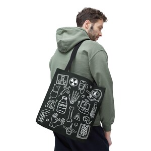 May include: Black tote bag with white medical-themed illustrations, including X-ray images, syringes, and a brain. The bag has a black shoulder strap and is carried by a person wearing a green hoodie.
