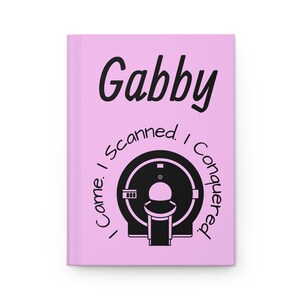 Op de afbeelding: Een lichtroze notitieboek met de naam "Gabby" in zwart schrift. Daaronder een zwarte illustratie van een medische scanner, omringd door de tekst "I came. I scanned. I conquered."