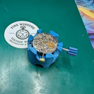 May include: A blue watch movement holder with a watch mechanism inside. The holder has a screw for adjustment. A white circular sticker with the text "FINE WINDING VINTAGE WATCH RESTORATION &" is next to the holder.