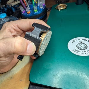 May include: A black watch repair tool holds a vintage watch movement. The watch movement has a gold-colored frame and a silver face. The background includes a green work surface, watch repair tools, and a sticker that reads "Fine Winding Vintage Watch Restoration & Service."