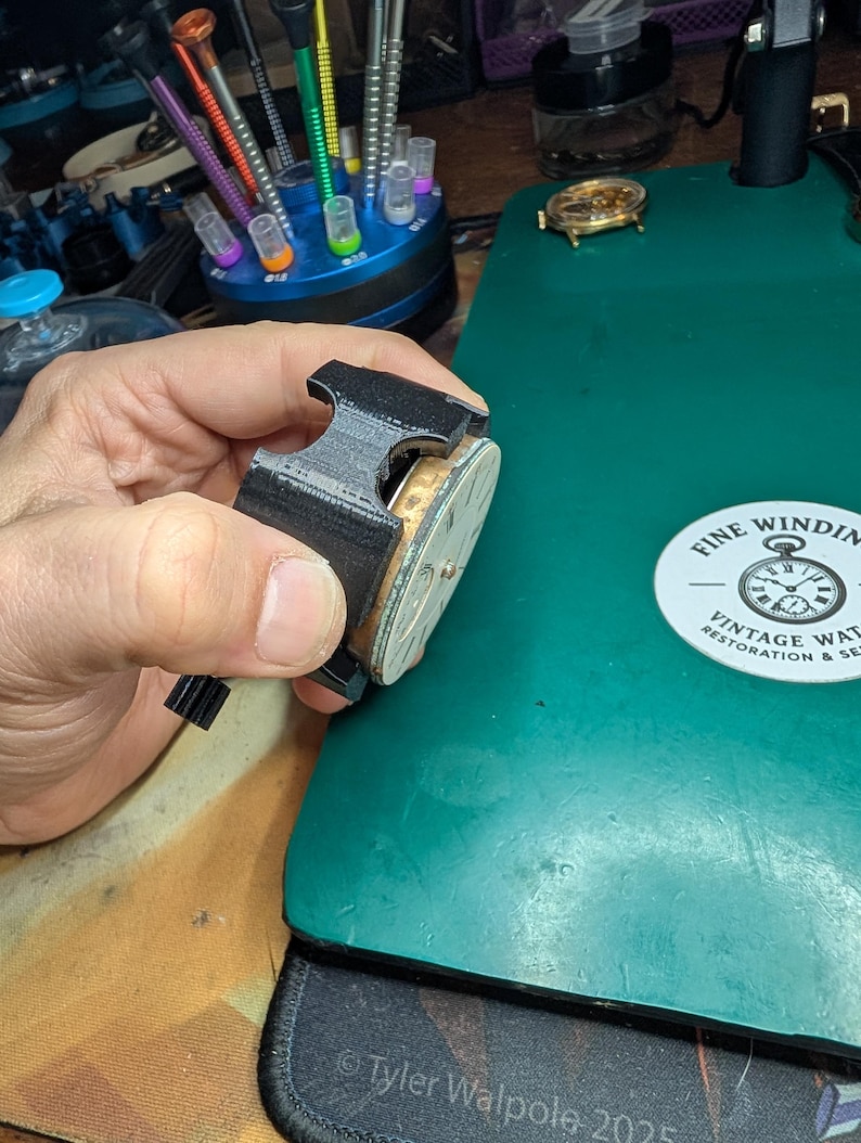 May include: A black watch repair tool holds a vintage watch movement. The watch movement has a gold-colored frame and a silver face. The background includes a green work surface, watch repair tools, and a sticker that reads "Fine Winding Vintage Watch Restoration & Service."