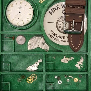 Puede incluir: Una bandeja organizadora verde llena de piezas de reloj, incluyendo esferas, engranajes y una correa de reloj de cuero marrón. La bandeja tiene compartimentos para varios componentes, con las palabras "FINE VINTAGE RESTORATION" en una esfera.