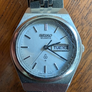Può includere: Un orologio da polso Seiko Quartz color argento con quadrante argento metallizzato, indici argentati e lancette nere. L'orologio presenta una finestrella giorno-data che mostra TUE 16, un bracciale a maglie in metallo e un logo.