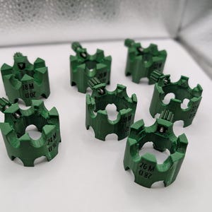 May include: A collection of seven green, gear-shaped components. Each component has a series of teeth around the outer edge and a small, cylindrical piece on top. The components are labeled with white text, including numbers and letters.