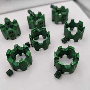 May include: A collection of seven green, gear-shaped components. Each component has a circular design with multiple teeth and a small, cylindrical extension. Some components have the text "18 M 0.02" printed on them. The objects are arranged on a white surface.