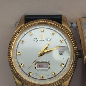 May include: A gold-tone automatic wristwatch with a white dial, gold hour markers, and gold hands. The dial features the text Dynamic Wide, Waterproof, 21 Jewels, and Japan. A date window displays the number 5 and Japanese characters.
