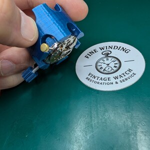 May include: A blue watch movement holder with a watch movement inside. The holder is being held by a hand. A white sticker with the text "FINE WINDING VINTAGE WATCH RESTORATION & SERVICE" is in the background.