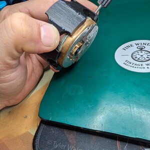 May include: A partially assembled watch movement with a black frame and brass components. The watch mechanism is held against a green surface, with a vintage watch restoration logo visible in the background.