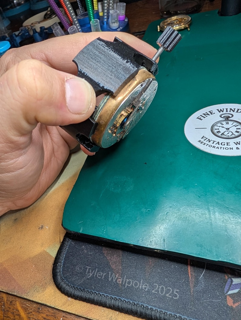 May include: A partially assembled watch movement with a black frame and brass components. The watch mechanism is held against a green surface, with a vintage watch restoration logo visible in the background.