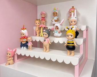 May include: A collection of twelve small, standing vinyl figurines displayed on a three-tiered white and pink scalloped shelf. The figures feature various costumes including a bee, a rabbit, a carousel, a mushroom, and a unicorn.