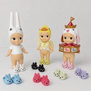 Op de afbeelding: Drie kleine figuurtjes met een getinte huid en grote ogen. Eén draagt een witte konijnenkap en shirt, één een gele eendenkap met jurk en vleugels, en één een carrousel-kap. Verschillende paren kleine, konijnvormige pantoffels zijn inbegrepen.