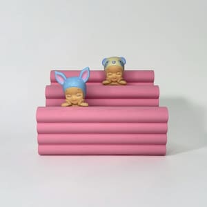 Może przedstawiać: R&oacute;żowy, warstwowy, dekoracyjny stojak wystawowy z dwiema małymi figurkami. Jedna figurka ma na sobie niebieską czapkę z uszami kr&oacute;lika, druga czapkę pandy. Figurki są beżowe, z zamkniętymi oczami i dłońmi pod brodą.