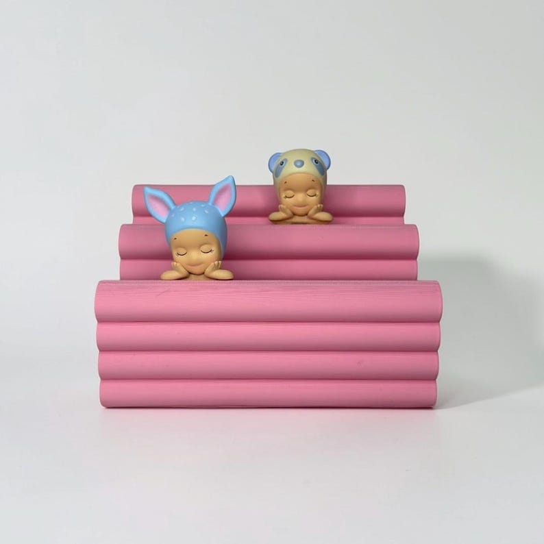 Może przedstawiać: R&oacute;żowy, warstwowy, dekoracyjny stojak wystawowy z dwiema małymi figurkami. Jedna figurka ma na sobie niebieską czapkę z uszami kr&oacute;lika, druga czapkę pandy. Figurki są beżowe, z zamkniętymi oczami i dłońmi pod brodą.