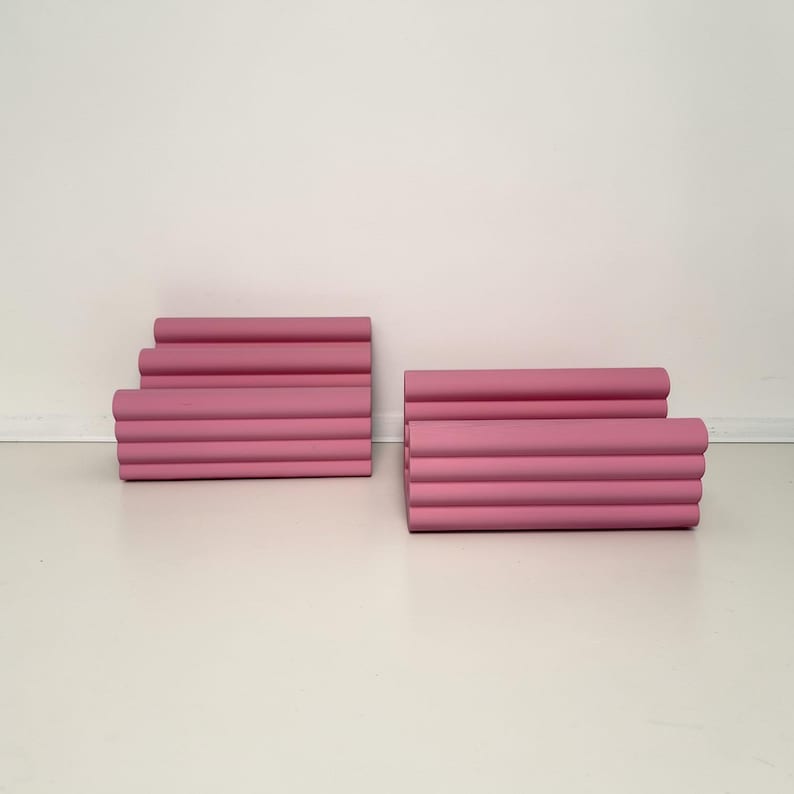 Może przedstawiać: Dwa zestawy r&oacute;żowych, cylindrycznych obiekt&oacute;w ułożonych w stos. Każdy zestaw składa się z wielu poziomych cylindr&oacute;w, tworząc efekt warstwowy. Obiekty mają jednolity, stonowany r&oacute;żowy kolor i są umieszczone na białej powierzchni.