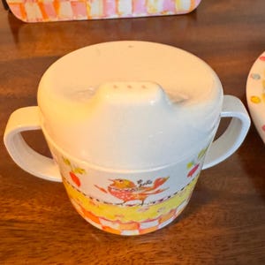 May include: A white sippy cup with two handles and a lid featuring a spout. The cup body is decorated with a colorful pattern of a bird, yellow bands, and an orange and white checkered base. The design includes small red and green accents.