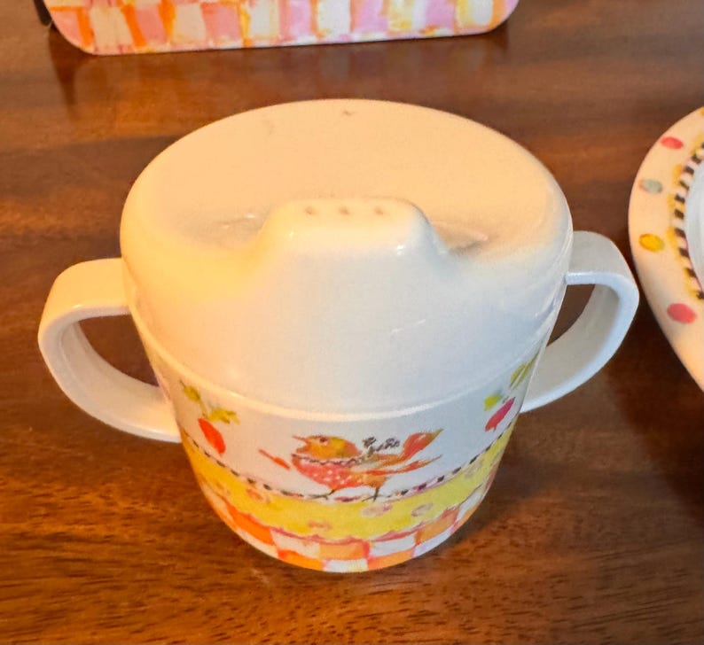 May include: A white sippy cup with two handles and a lid featuring a spout. The cup body is decorated with a colorful pattern of a bird, yellow bands, and an orange and white checkered base. The design includes small red and green accents.