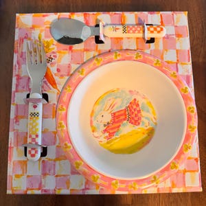 May include: A white ceramic bowl with a pink rim featuring a painted rabbit in a red and white striped shirt, accompanied by a matching metal fork and spoon with patterned handles, all presented on a pink and orange checkered display card.