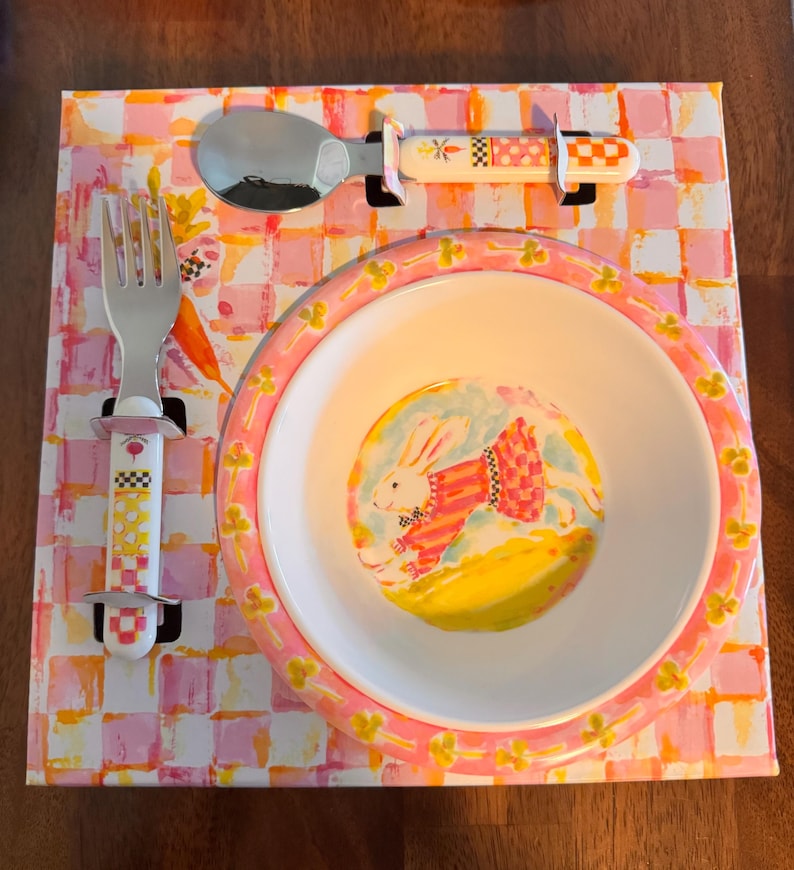 May include: A white ceramic bowl with a pink rim featuring a painted rabbit in a red and white striped shirt, accompanied by a matching metal fork and spoon with patterned handles, all presented on a pink and orange checkered display card.