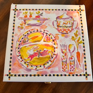 May include: A square white box featuring a colorful watercolor illustration of a divided plate with a rabbit, a bowl of carrots, a lidded cup, a fork, and a spoon. The artwork is framed by a black and white checkered border on a pink background.