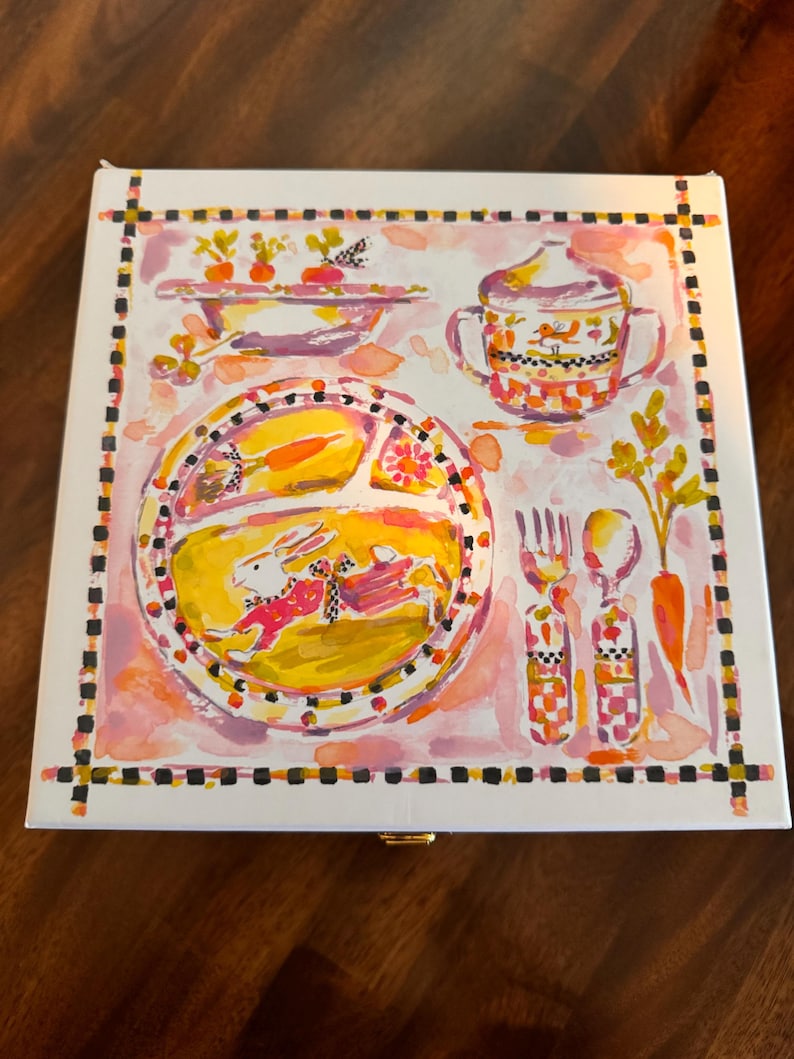 May include: A square white box featuring a colorful watercolor illustration of a divided plate with a rabbit, a bowl of carrots, a lidded cup, a fork, and a spoon. The artwork is framed by a black and white checkered border on a pink background.