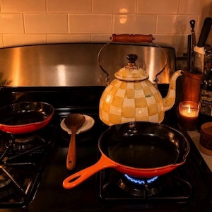 May include: A black stovetop featuring two orange cast iron skillets, one of which is placed over a blue gas flame. A yellow and white checkered tea kettle with a wooden handle sits behind the pans. A wooden spoon, two candles, and a bottle are nearby.