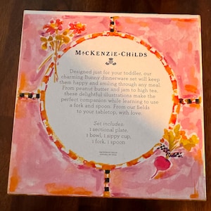 May include: A square product box for a MacKenzie-Childs Bunny dinnerware set. The box features a pink watercolor background with floral and vegetable illustrations. The center text lists the set contents: 1 sectional plate, 1 bowl, 1 sippy cup, 1 fork, and 1 spoon, along with brand information and descriptive text.