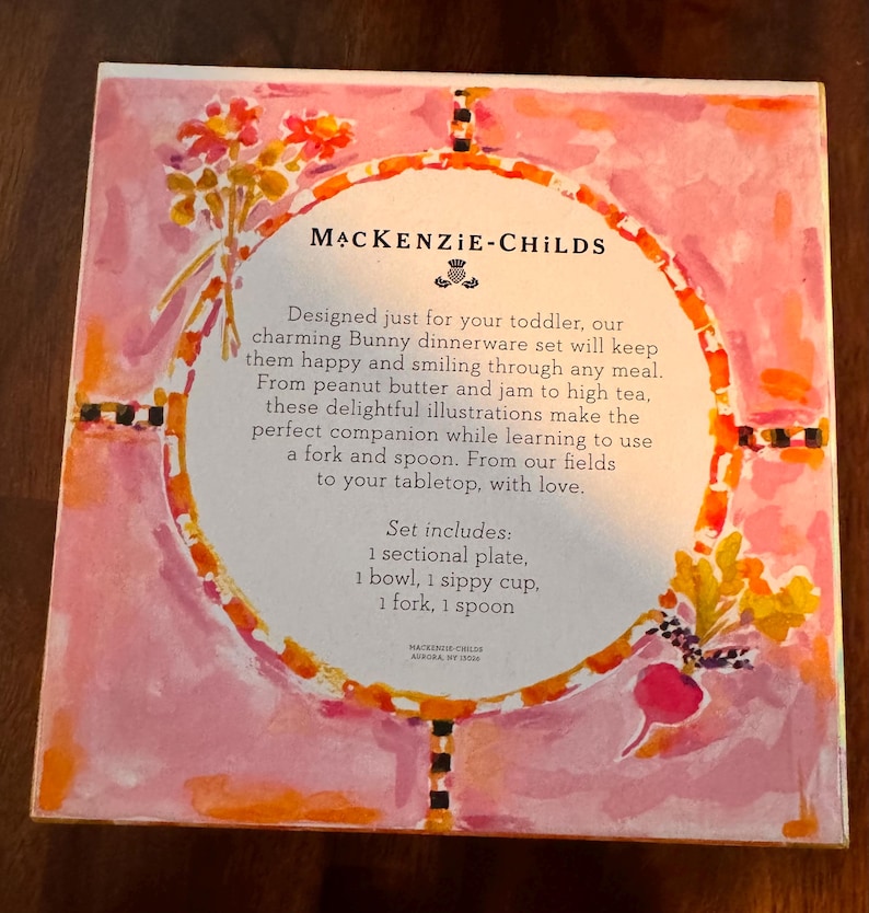May include: A square product box for a MacKenzie-Childs Bunny dinnerware set. The box features a pink watercolor background with floral and vegetable illustrations. The center text lists the set contents: 1 sectional plate, 1 bowl, 1 sippy cup, 1 fork, and 1 spoon, along with brand information and descriptive text.