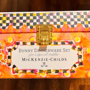 May include: A rectangular box with a gold-tone metal latch, featuring a vibrant orange, yellow, and pink watercolor-style pattern with a black and white checkered border. The label reads: BUNNY DINNERWARE SET for a special toddler MACKENZIE-CHILDS.