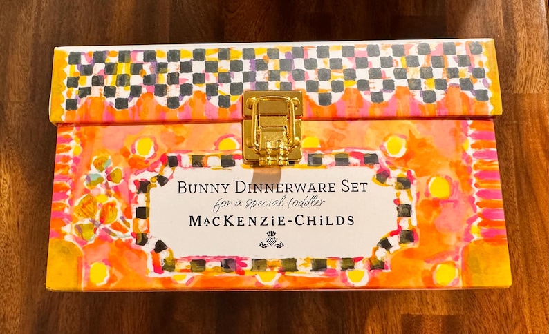 May include: A rectangular box with a gold-tone metal latch, featuring a vibrant orange, yellow, and pink watercolor-style pattern with a black and white checkered border. The label reads: BUNNY DINNERWARE SET for a special toddler MACKENZIE-CHILDS.